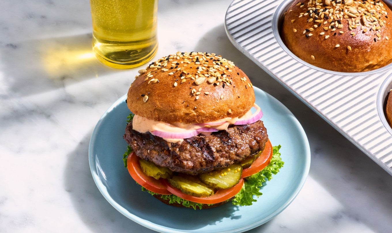 A Whole Wheat Hamburger Bun filled with a hamburger, lettuce, pickles, tomato, and onion on a marble countertop.